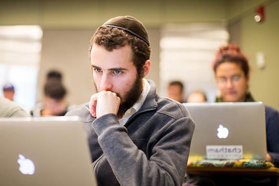 Photo of a male student in a yarmulke, 坐在冰球突破app的演讲厅里，看着他的电脑
