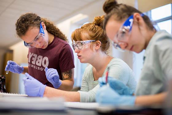 图为三名冰球突破app的女学生戴着护目镜和手套在实验室工作