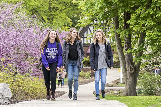 春季，冰球突破app的三名女学生在冰球突破mg登录网址步行去上课, 有绿色的树和五颜六色的花. 